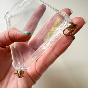 May include: A clear phone case with a gold ring and a gold clasp attached to the bottom. The case is made of a flexible material and has a raised lip around the edges to protect the screen.