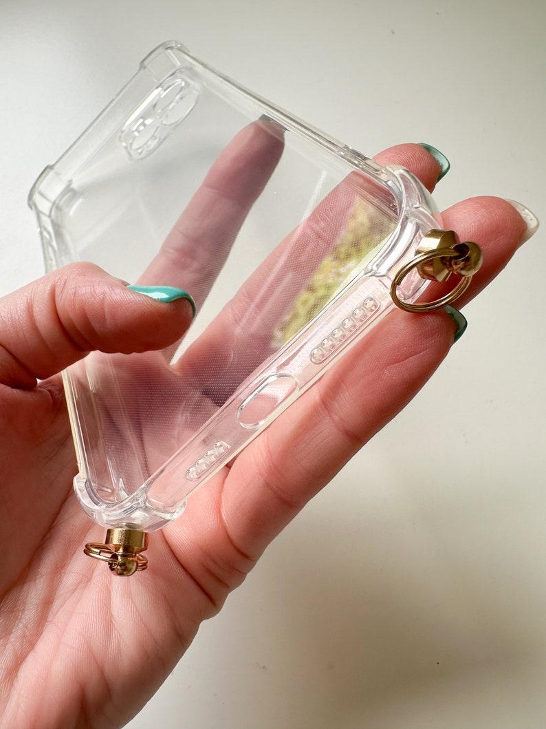 May include: A clear phone case with a gold ring and a gold clasp attached to the bottom. The case is made of a flexible material and has a raised lip around the edges to protect the screen.