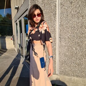 May include: A woman wearing a black floral crop top, beige pants, and sunglasses. She is carrying a blue phone case attached to a gold chain.