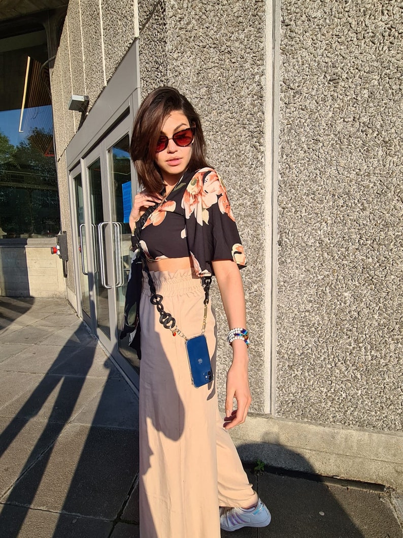 May include: A woman wearing a black floral crop top, beige pants, and sunglasses. She is carrying a blue phone case attached to a gold chain.