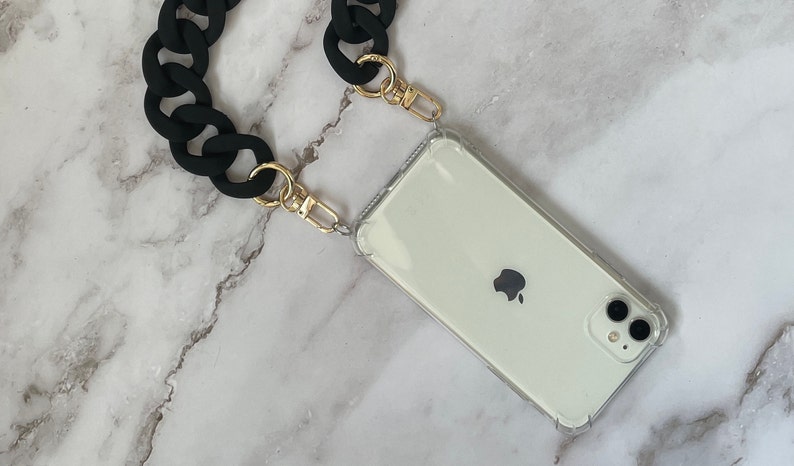 May include: A black chain phone strap with gold clasps attached to a clear phone case with a white phone inside.