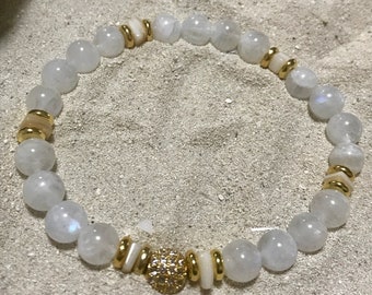 May include: A beaded bracelet featuring round, translucent white beads interspersed with gold-colored spacers and a central gold-colored bead with embedded crystals. The bracelet is displayed on a sandy surface.