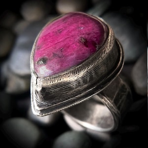 以下が含まれることがあります： 洋梨型の深いマゼンタ色の宝石をあしらった指輪。質感のある酸化銀のバンドにセットされています。金属製の台座には目に見える縦の筋が入っており、磨かれた不透明な石の周囲には層状の階段状のデザインが施されています。