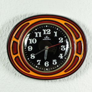 Puede incluir: Un reloj redondo de cerámica naranja y rojo con números y manecillas negros. La esfera del reloj dice "Meissner Anker" y tiene un fondo negro.