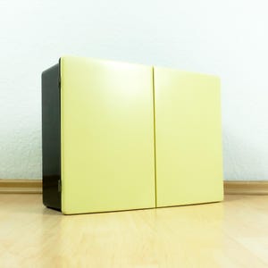 Könnte beinhalten: Ein zweitüriger Schrank mit schwarzem Rahmen und hellgelben Türen. Der Schrank ist rechteckig und steht auf einem hellen Holzboden. Die Türen sind bündig und haben eine vertikale Naht.