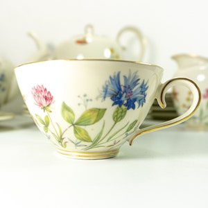 Könnte beinhalten: Eine weiße Teetasse mit einem Blumendesign mit blauen, rosa und grünen Blumen. Die Tasse hat einen goldenen Rand und einen zarten Henkel.