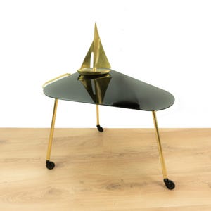 Könnte beinhalten: Ein moderner Beistelltisch mit einer schwarzen, dreieckigen Tischplatte und goldfarbenen Beinen. Auf der Tischoberfläche befindet sich eine kleine Segelboot-Skulptur aus Messing. Der Tisch hat drei Beine und kleine Rollen.