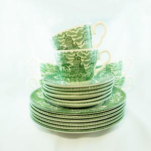 May include: A stack of green and white teacups and saucers. The cups and saucers have a green landscape design. The cups are stacked on top of the saucers. The teacups have white handles.