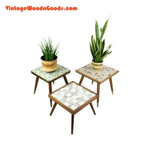 May include: Three mid-century modern plant stands with tiled tops. The stands are made of wood and have a brown finish. The tiles are in a geometric pattern and are in shades of green, brown, and white.