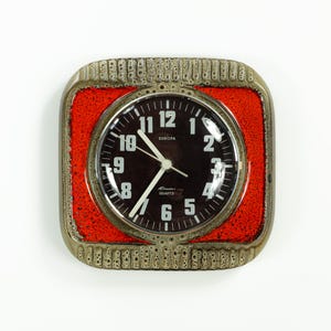 May include: A vintage Europa quartz clock. The clock has a black face with white numerals and hands. It is set in a square frame with a red centre and a textured, brown border. The clock face reads "Europa Quartz".