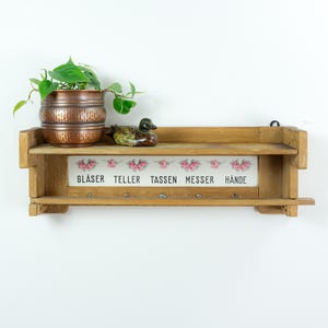 Könnte beinhalten: Rustikales Holzregal mit zwei Ebenen, mit einem kupferfarbenen Topf mit einer grünen Pflanze, einer Keramik-Entenfigur und einer dekorativen Tafel mit floralen Akzenten und deutschem Text. Unter dem Regal befinden sich Haken.
