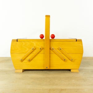Könnte beinhalten: Ein Nähkasten aus hellem Holz mit einem ausziehbaren Akkordeon-Mechanismus und einem zentralen vertikalen Griff. Der Kasten verfügt über zwei kleine, runde, leuchtend rote Knöpfe auf dem Deckel und steht auf vier kleinen Holzfüßen.