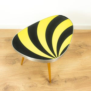 Könnte beinhalten: Ein Beistelltisch im Retro-Stil mit einem schwarz-gelb gestreiften Design auf der ovalen Tischplatte. Der Tisch hat einen silbernen Rand und drei Holzbeine. Der Tisch ist ca. 61 cm lang, 41 cm breit und 46 cm hoch.