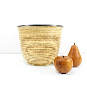 Könnte beinhalten: Ein brauner und beigefarbener Keramiktopf mit schwarzem Rand. Der Topf hat ein gestreiftes Muster und steht auf einer weißen Oberfläche mit zwei Holzbirnen daneben.