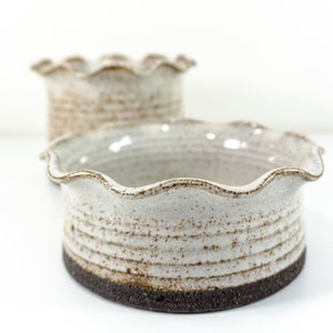 Könnte beinhalten: Zwei weiße Keramikschalen mit einem gewellten Rand und einer braunen Glasur. Die Schalen sind handgefertigt und haben einen rustikalen Look.