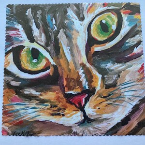 May include: A square art print featuring a close-up painting of a tabby cat face with prominent green eyes, a pink nose, and textured brushstrokes in shades of brown, tan, black, and white. The edges of the print have a decorative scalloped border.