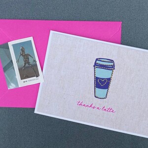 May include: A greeting card with a coffee cup design and the words "thanks a latte" in pink. The card is on a textured background, next to a pink envelope and a stamp with a robot illustration.