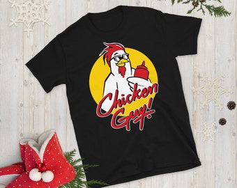 Può includere: Maglietta nera con un'immagine di un pollo dei cartoni animati che indossa occhiali da sole e tiene una bottiglia di ketchup. Il testo "Chicken Guy!" è scritto in corsivo rosso e bianco.