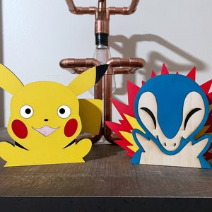 Op de afbeelding: Twee houten beeldjes van Pokémon-personages. De eerste is een gele Pikachu met rode wangen en zwarte oren. De tweede is een blauwe Charizard met rode vlammen en een gele onderkant.