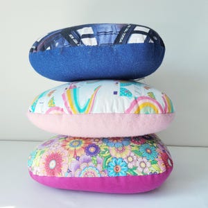 May include: Three stacked, oval-shaped pillows. The top pillow is blue with a pattern of a police box. The middle pillow has a white background with rainbows and pastel colors. The bottom pillow has a floral pattern with pink and purple accents.