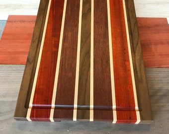 May include: A rectangular wooden cutting board with a natural wood finish. The board has a raised edge and is made of multiple types of wood, including walnut, maple, and cherry. The wood is arranged in a striped pattern.