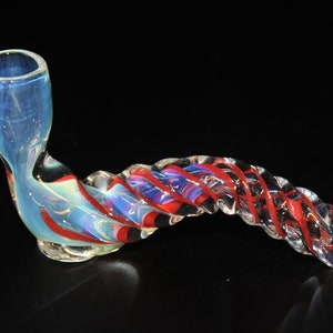 May include: A clear glass pipe with a blue bowl and a twisted stem. The stem features red and blue spiral accents. The pipe is designed with a curved shape and sits on a black surface.