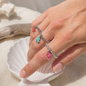Può includere: Due anelli d'argento con perline e ciondoli di creature marine colorate. Un anello presenta una stella marina rossa, l'altro ha un pesce blu e un cavalluccio marino rosa. Gli anelli sono mostrati su una mano vicino a una conchiglia bianca.