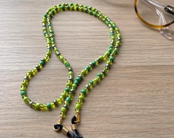 Könnte beinhalten: Eine grün-graue Brillenkette aus Perlen. Die Kette besteht aus kleinen, runden Perlen und hat goldfarbene Schlaufen zur Befestigung an Brillen. Die Kette liegt auf einer Holzoberfläche, im Hintergrund eine Brille.
