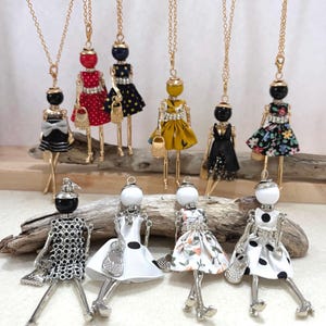 May include: A collection of doll pendant necklaces with gold chains. The dolls have various dresses in colors like black, red, yellow, and floral prints. Some dolls have small purses. The dolls are made of metal and beads.