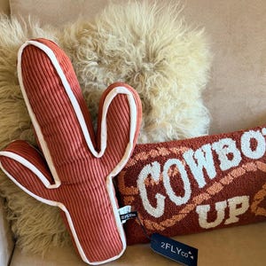 Könnte beinhalten: Ein dekoratives Kissen in Kaktusform und ein rechteckiges Kissen mit dem Text "COWBOY UP" in Weiß und Orange. Das Kaktuskissen ist rostfarben mit weißer Umrandung. Maße in Zentimetern.