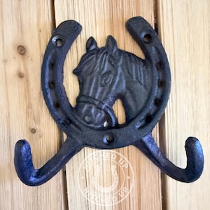 May include: A black cast iron wall hook featuring a horseshoe shape with a horse head relief in the center. The design includes two curved hooks at the bottom and pre-drilled holes for mounting. A circular logo with the text STITCHED UP is visible.