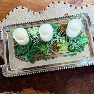 May include: A rectangular silver tray with beaded edges and handles, holding three white pillar candles surrounded by faux succulents. The tray sits on a cowhide-style placemat with a studded border. The text "HITCHED UP" is visible.