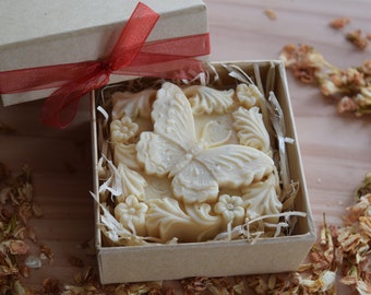 Puede incluir: Una pastilla de jabón blanca con un diseño de mariposa en una caja marrón con una cinta roja. El jabón está rodeado de papel triturado.