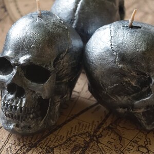 May include: Three silver skull-shaped candles with visible details of the skull's structure. Each candle has a wick at the top. The candles are placed on a vintage map, creating a gothic or Halloween theme.