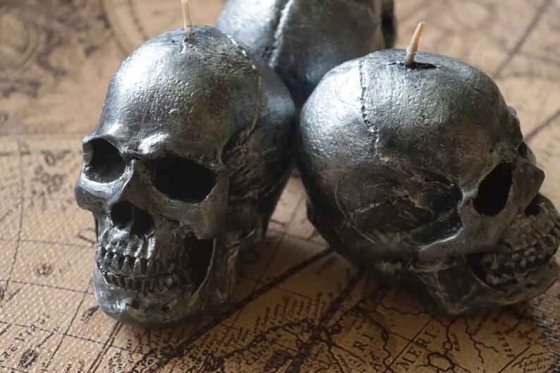 May include: Three silver skull-shaped candles with visible details of the skull's structure. Each candle has a wick at the top. The candles are placed on a vintage map, creating a gothic or Halloween theme.