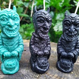 Puede incluir: Tres velas con temática de Halloween con forma de demonio sosteniendo un caldero. Las velas son verdes, negras y negras.