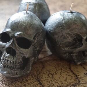 May include: Three silver-colored skull candles with wicks. The candles are detailed with realistic skull features, including eye sockets, a nose cavity, and teeth. The candles are placed on a map.