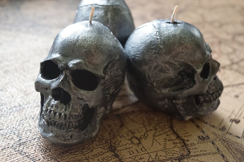 May include: Three silver-colored skull candles with wicks. The candles are detailed with realistic skull features, including eye sockets, a nose cavity, and teeth. The candles are placed on a map.