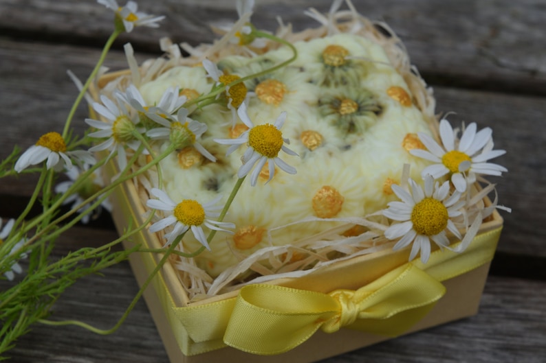 May include: A yellow gift box with a yellow bow filled with a white crocheted soap with green and gold accents. The box is surrounded by white daisies.