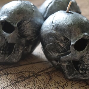 May include: Three silver-colored skull candles with a textured finish. The candles are placed on a vintage map, creating a gothic or Halloween theme. Each skull has detailed facial features and a wick.