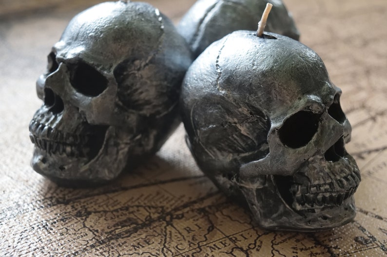 May include: Three silver-colored skull candles with a textured finish. The candles are placed on a vintage map, creating a gothic or Halloween theme. Each skull has detailed facial features and a wick.