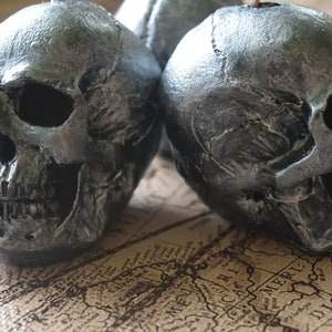 May include: Close-up of two silver-painted skull figurines. The skulls have detailed features, including eye sockets, nasal cavities, and teeth. The skulls are placed on a vintage map background, creating a gothic or Halloween theme.