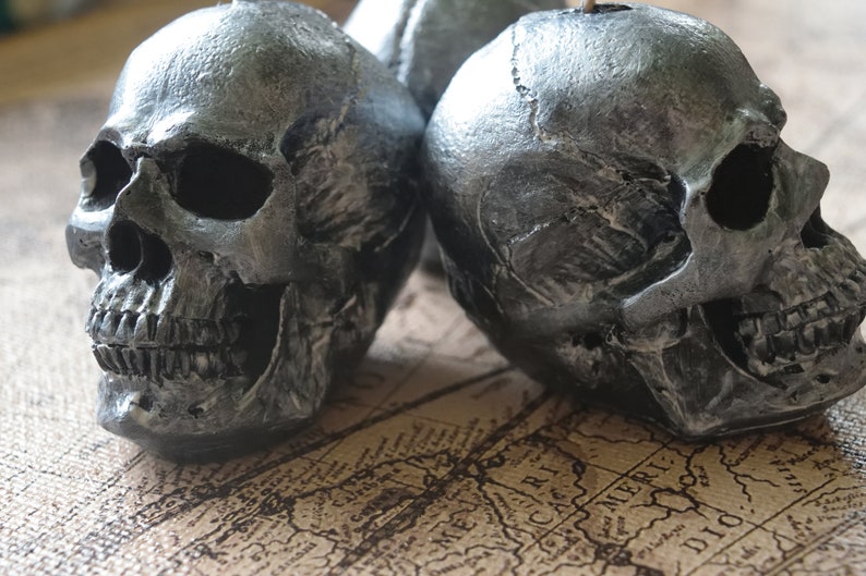May include: Close-up of two silver-painted skull figurines. The skulls have detailed features, including eye sockets, nasal cavities, and teeth. The skulls are placed on a vintage map background, creating a gothic or Halloween theme.