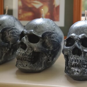 May include: Three skull-shaped candles with a metallic gray finish. Each candle has a wick at the top. The candles are arranged in a row, showcasing their detailed skeletal features, perfect for Halloween decor.