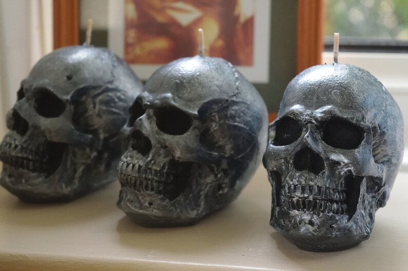 May include: Three skull-shaped candles with a metallic gray finish. Each candle has a wick at the top. The candles are arranged in a row, showcasing their detailed skeletal features, perfect for Halloween decor.