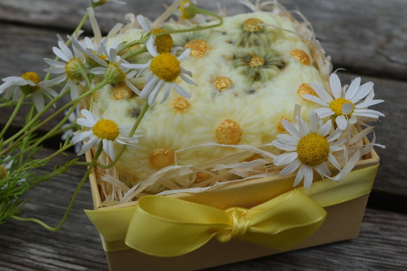 May include: A yellow gift box with a yellow ribbon bow, filled with a white soap bar decorated with daisies and gold accents. The box is surrounded by white daisies.