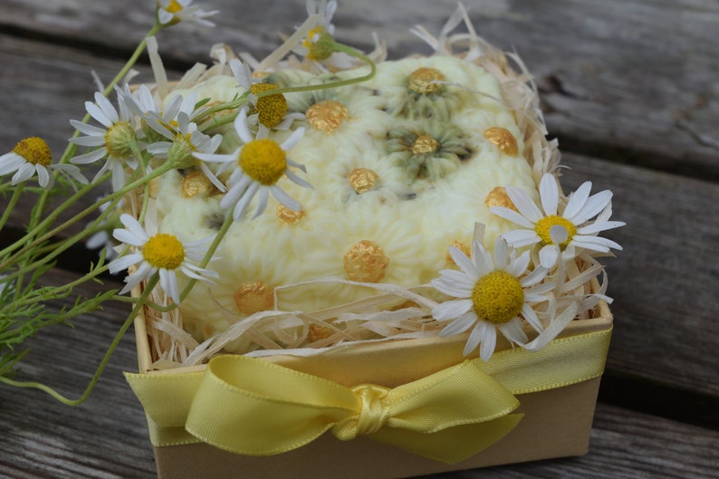 May include: A yellow gift box with a yellow bow and a white soap bar decorated with chamomile flowers. The box is filled with shredded paper and surrounded by chamomile flowers.