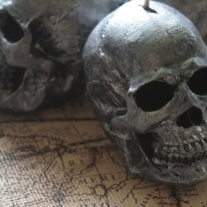 May include: Three gray skull-shaped candles with a textured surface. The candles have a wick at the top and are placed on a map-like background. The skulls have detailed facial features.