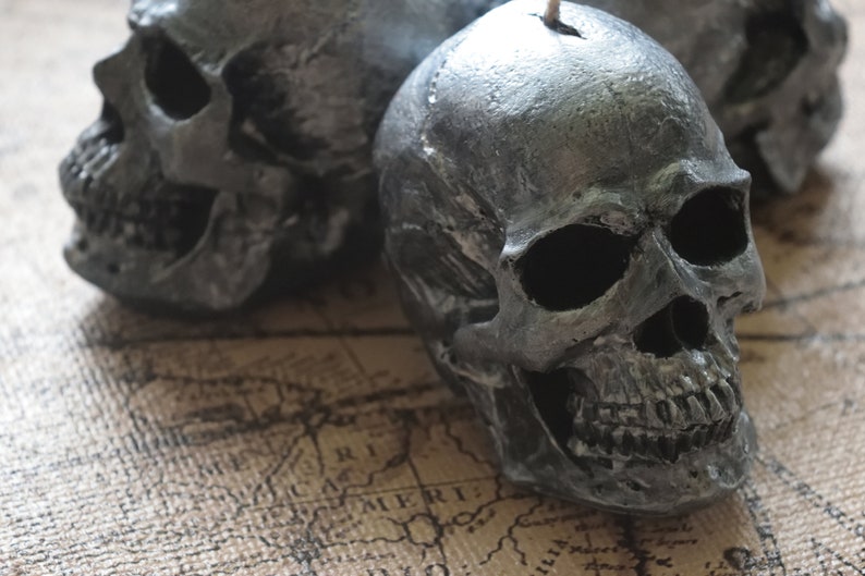 May include: Three gray skull-shaped candles with a textured surface. The candles have a wick at the top and are placed on a map-like background. The skulls have detailed facial features.