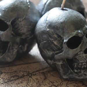 May include: Close-up of three gray skull-shaped candles. The candles have a textured, metallic finish and are arranged on a map-like surface. The candles are a decorative item, perfect for Halloween or gothic decor.